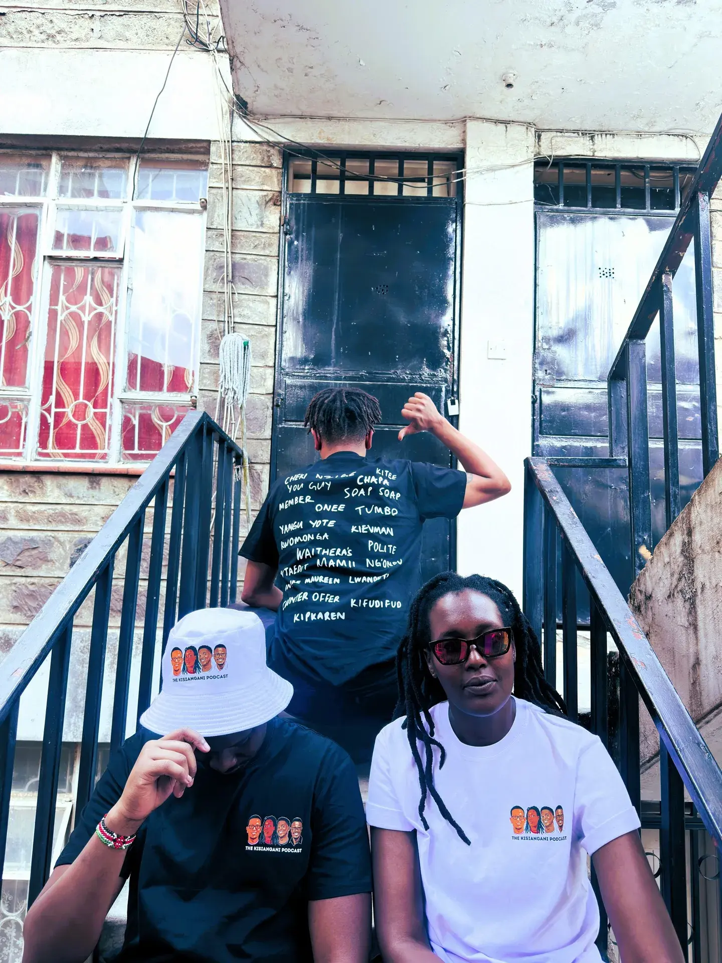 Three people on a stairway in Kisiangani Podcast tees and a bucket hat
